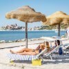 Persone su lettini sotto ombrelloni di paglia, vista su mare e borgo in lontananza.